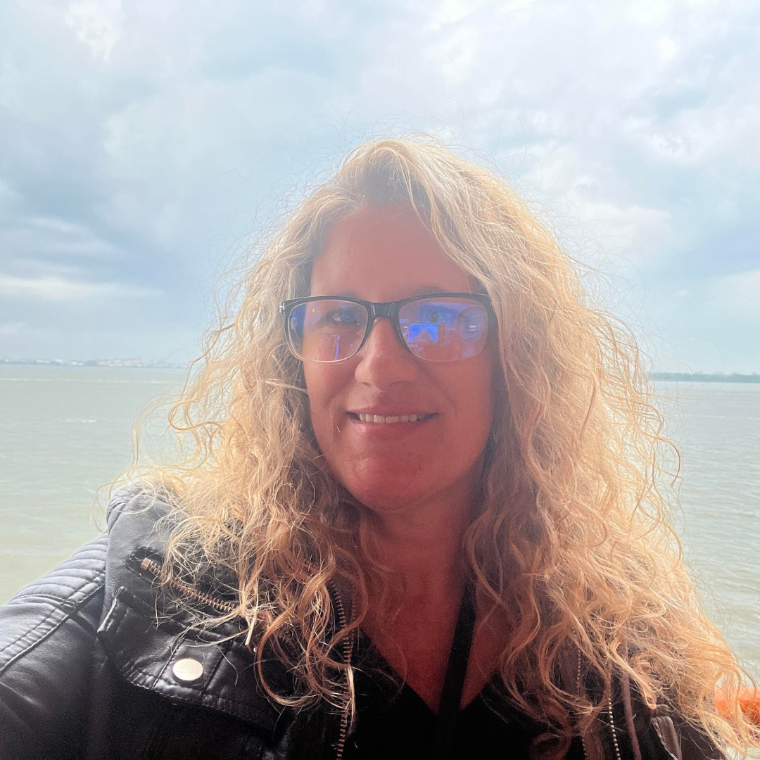 Photo of Pastor Jill Beck with the ocean in the background. She is smiling and has curly hair and glasses.